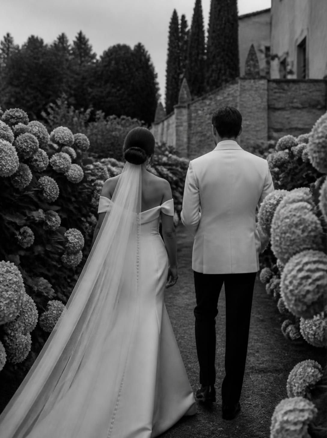 Fotografia in bianco e nero: gli sposi, di spalle, camminano lungo un sentiero tra le ortensie