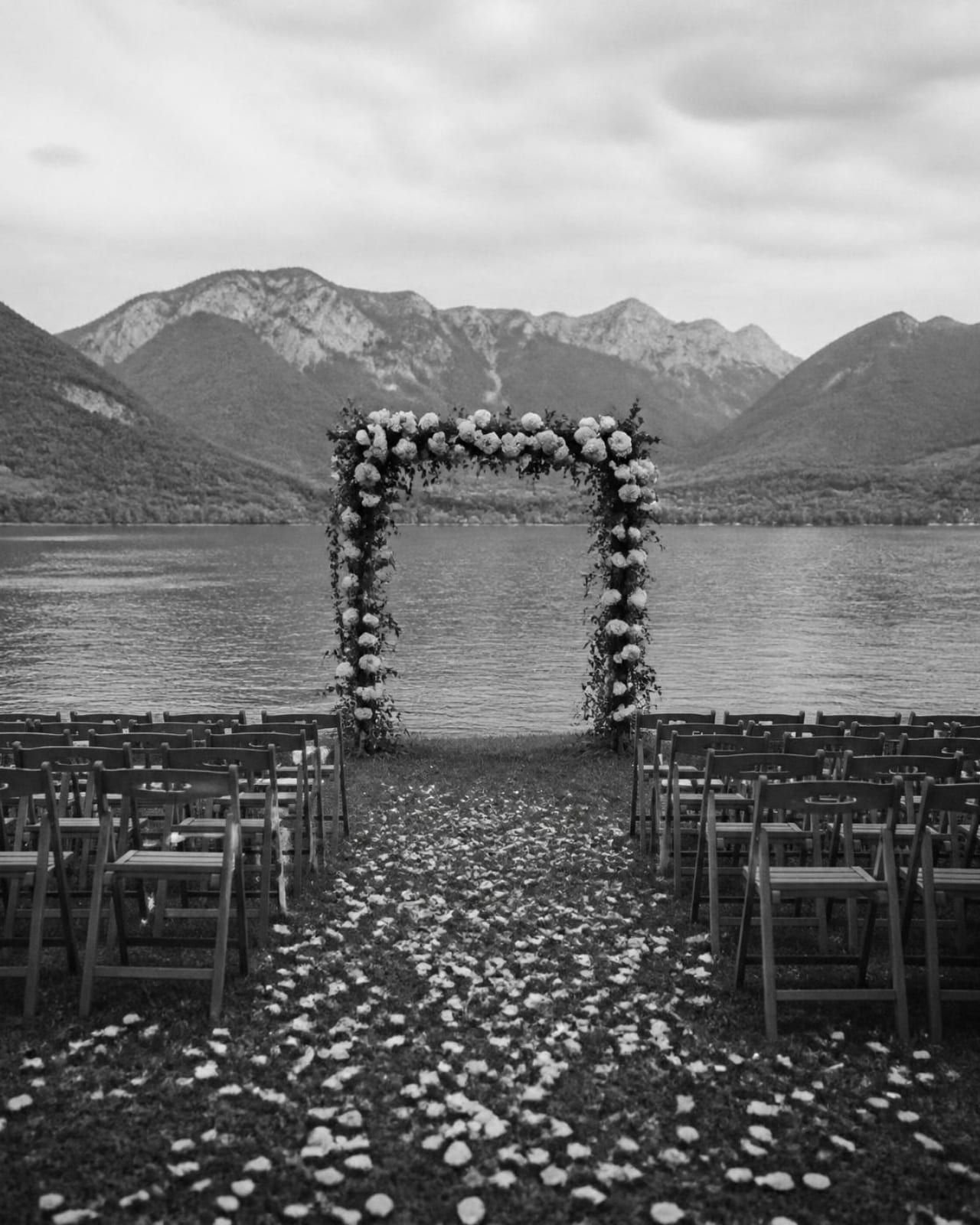 Sillas de madera en filas sobre un prado junto al lago, con un pasillo de pétalos y un arco de rosas blancas, en blanco y negro