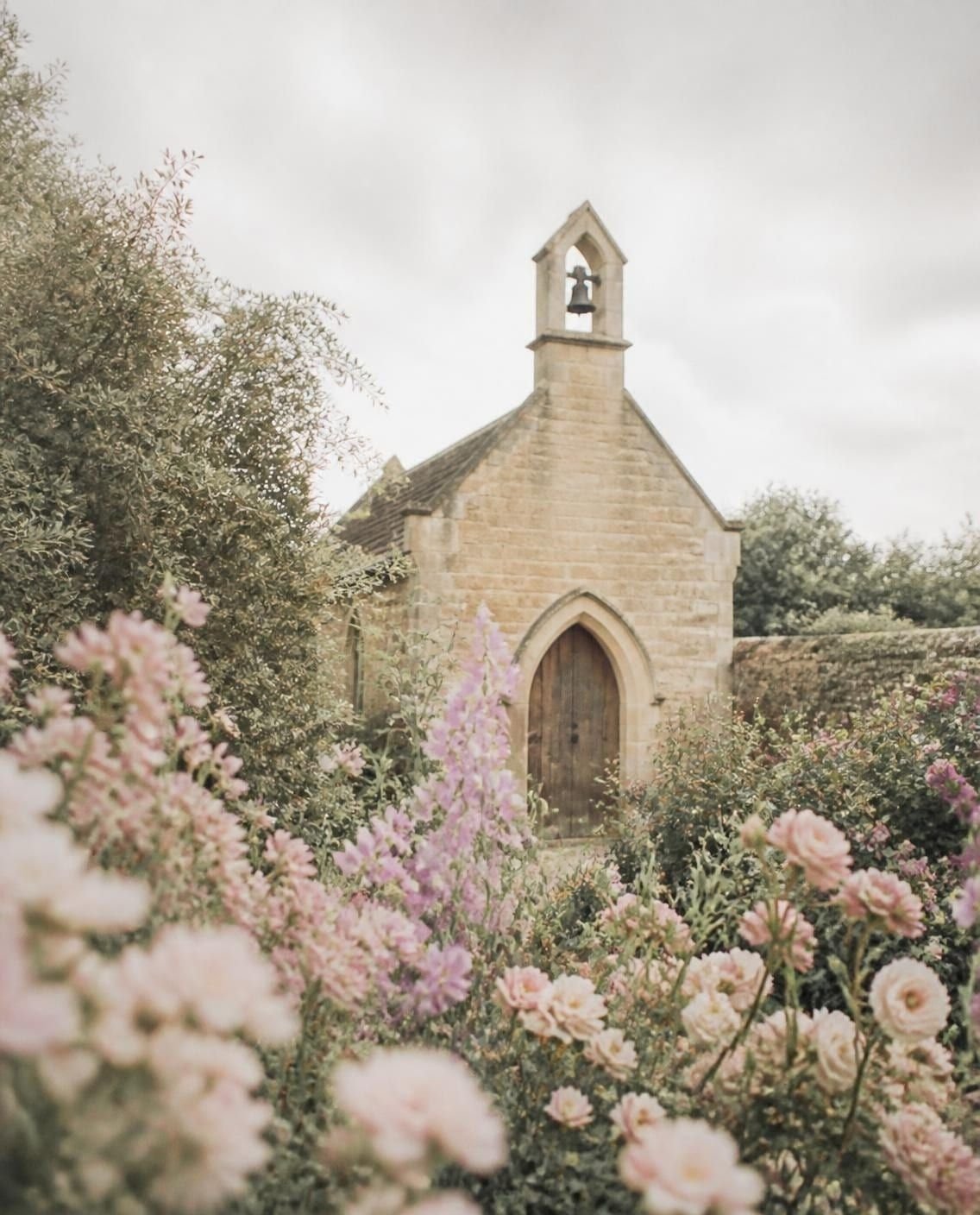 Capilla de piedra color miel con espadaña en un jardín amurallado, con rosas de jardín y flores lilas en primer plano