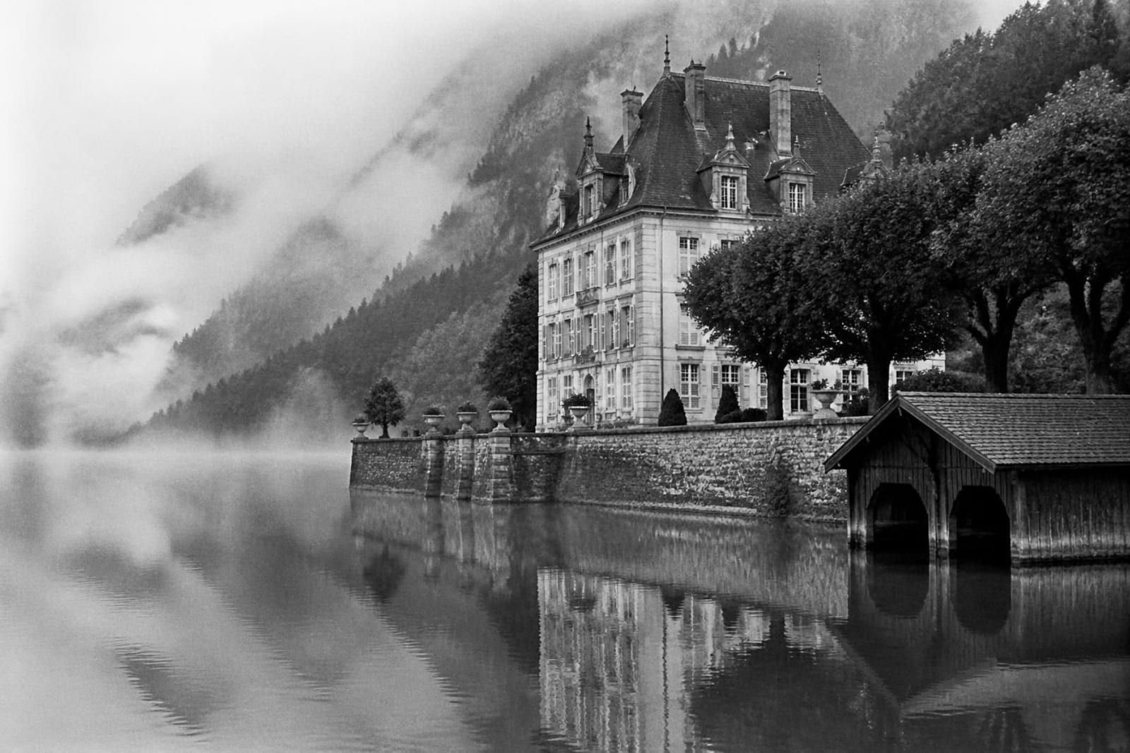 Pequeño château con tejados de pizarra a orillas de un lago de montaña, con una caseta de barcas y bruma sobre el agua, en blanco y negro