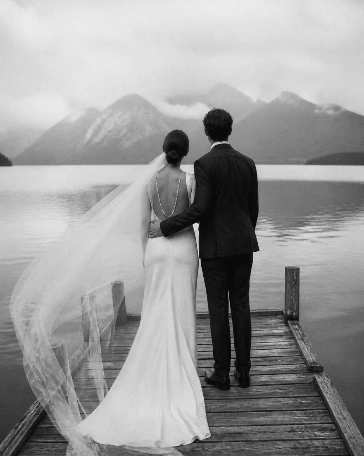Retrato en blanco y negro de los novios, de espaldas, en un embarcadero de madera sobre el lago, con el velo al viento y las montañas entre la bruma