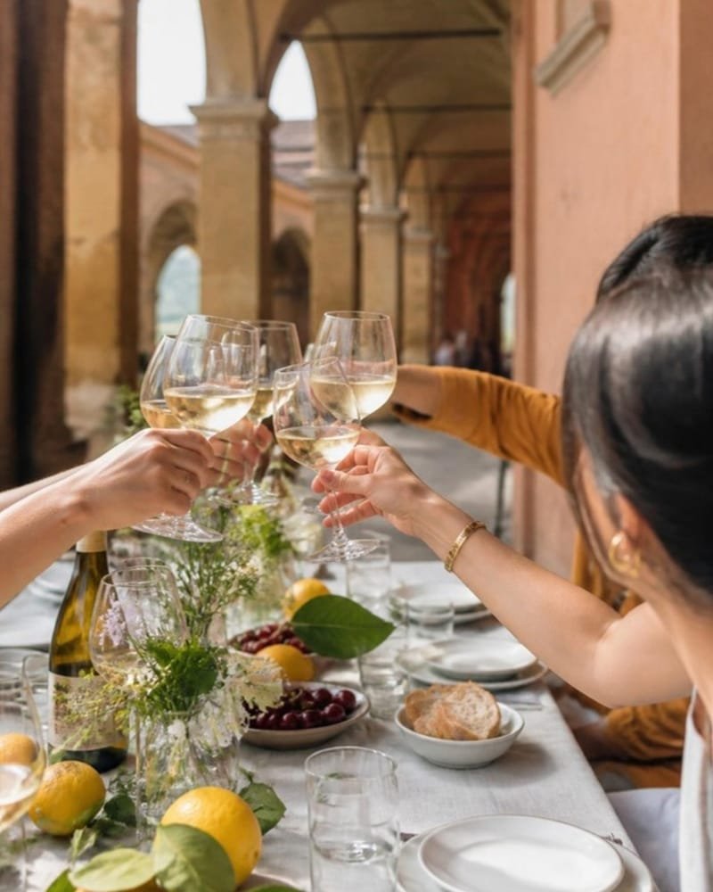 Amigos que brindan con vino blanco en una mesa larga bajo los pórticos, a la luz de la tarde