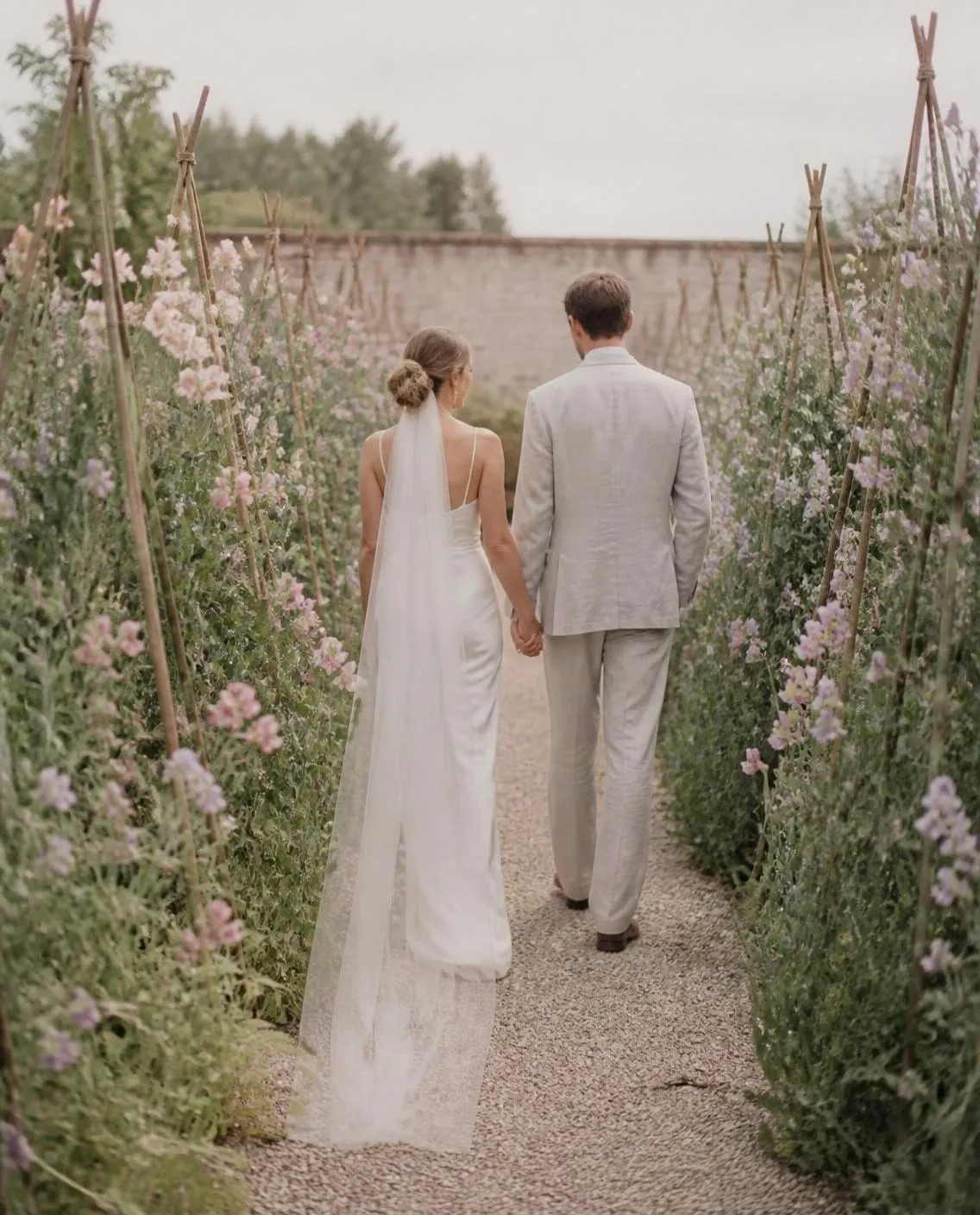 Pareja de espaldas, de la mano, en un camino de grava entre tutores de guisantes de olor rosas y lilas