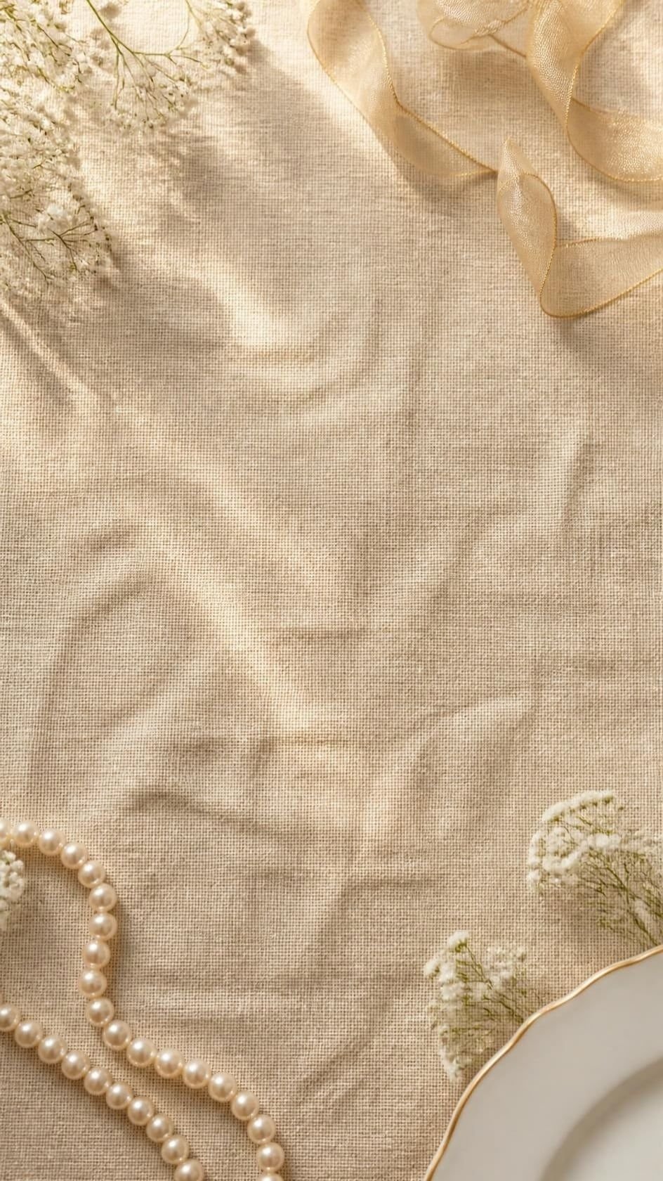 Tovaglia di lino champagne vista dall’alto, con un filo di perle, un nastro di organza dorata, rametti di gipsofila bianca e il bordo di un piatto di porcellana con il filo d’oro